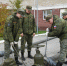 Четыре человека в военной форме занимаются соединением водопроводных труб. На земле лежат три больших вентиля, соединенных трубами. Один человек использует рычаг для затягивания соединения. Все четверо одеты в камуфляжную форму и черные ботинки. На рукава - ЮГУ